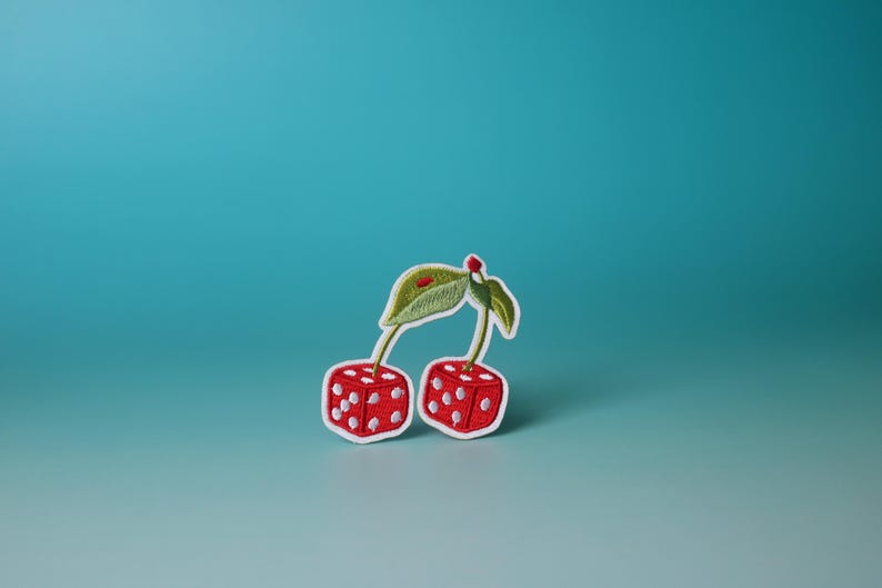 Puede incluir: Un parche bordado con dos cerezas, cada una con forma de dado, con puntos rojos y blancos. Los tallos y las hojas son verdes.