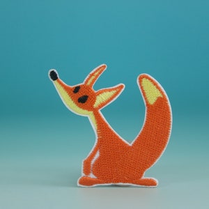 Peut inclure: Patch brodé en forme de renard orange avec des yeux noirs et une queue jaune. Le renard est debout sur ses pattes arrière, la tête levée.