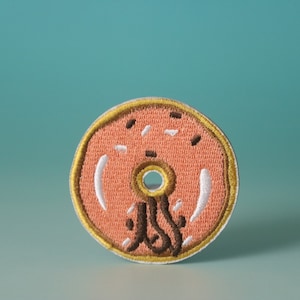 May include: A pink embroidered donut patch with brown chocolate icing and white sprinkles. The donut has a yellow outline.