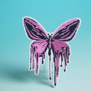 Peut inclure: Écusson papillon brodé avec un design unique. Le papillon présente des ailes roses avec des détails noirs et un effet dégoulinant. L'écusson a une bordure blanche et est posé sur un fond bleu dégradé.