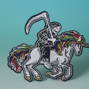 Puede incluir: Parche bordado con una figura esquelética negra montando un unicornio blanco con una crin y un cuerno arcoíris. La figura sostiene una guadaña.