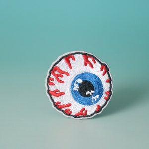 May include: A white, red and blue embroidered eye patch with a black pupil. The eye is surrounded by a red, dripping blood effect.