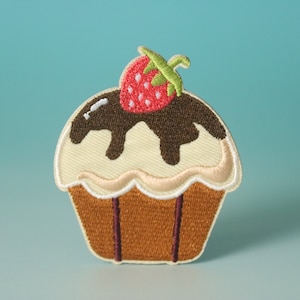 Può includere: Un cupcake marrone e bianco con glassa al cioccolato e una fragola rossa in cima. Il cupcake è ricamato su un tessuto bianco.