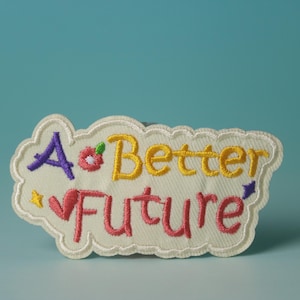 Może przedstawiać: Biała naszywka haftowana z tekstem "A Better Future" w kolorze żółtym, różowym, fioletowym i zielonym. Naszywka ma wzór kwiatu i serca.