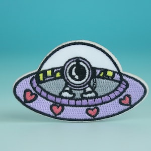 May include: Purple and white embroidered patch of a cartoon alien in a flying saucer with hearts around the edge.