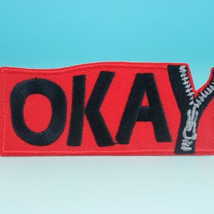 May include: Red fabric patch with the word "OKAY" embroidered in black with a silver zipper sewn into the right side of the patch.