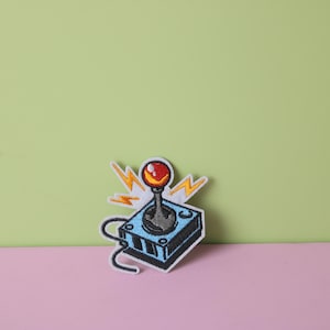 May include: An embroidered patch depicting a retro video game joystick in blue and black, with a red button and yellow lightning bolts.