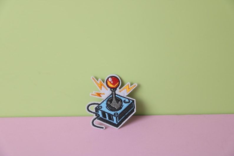 May include: An embroidered patch depicting a retro video game joystick in blue and black, with a red button and yellow lightning bolts.