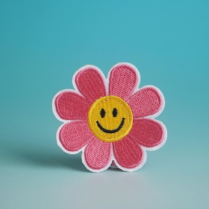 以下が含まれることがあります： 青い背景に、黄色い笑顔が刺繍されたピンクの花。