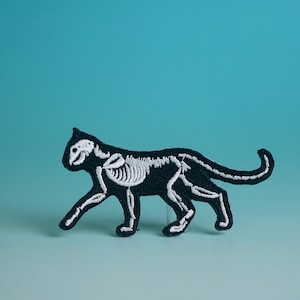 May include: Black embroidered patch of a cat skeleton with white stitching. The cat is walking with its tail curled up.