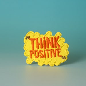 Könnte beinhalten: Gelber gestickter Aufnäher mit dem Text "THINK POSITIVE" in orangefarbenem Garn.