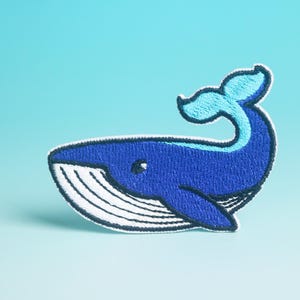 May include: Embroidered patch featuring a blue whale design with a light blue tail and accents. The whale has a white belly and is outlined in black. The patch is on a light blue background.