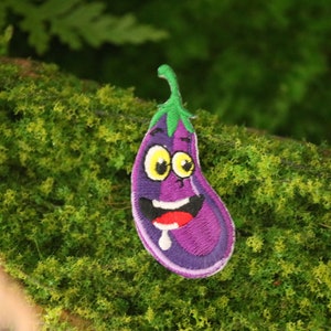 May include: A purple eggplant with a smiling face embroidered on a green background. The eggplant has a green stem and white eyes.