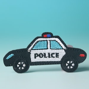 Puede incluir: Un parche bordado de un coche de policía blanco y negro con una luz azul en la parte superior. El coche tiene la palabra "POLICE" escrita en el lateral.