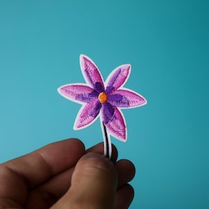 May include: A purple and pink embroidered flower patch with an orange center. The flower has five petals and a stem.