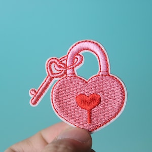 Puede incluir: Candado rosa en forma de corazón con una llave adjunta. El candado tiene un corazón rojo más pequeño en el centro.