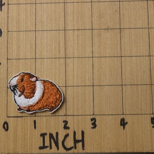 K&ouml;nnte beinhalten: Gestickter B&uuml;gel-Aufn&auml;her mit einem braunen und wei&szlig;en Meerschweinchen, etwa 2,5 cm breit, auf einer karierten Holzoberfl&auml;che.