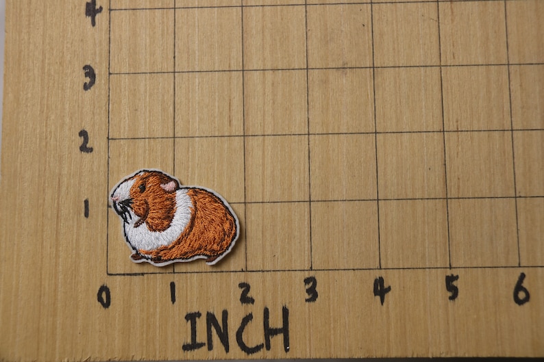 K&ouml;nnte beinhalten: Gestickter B&uuml;gel-Aufn&auml;her mit einem braunen und wei&szlig;en Meerschweinchen, etwa 2,5 cm breit, auf einer karierten Holzoberfl&auml;che.