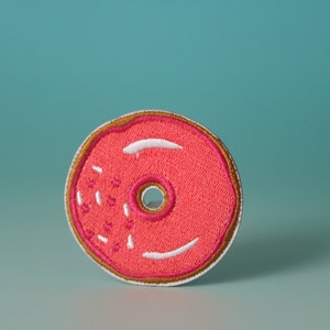 May include: A pink embroidered donut patch with white frosting and sprinkles. The donut has a brown outline.
