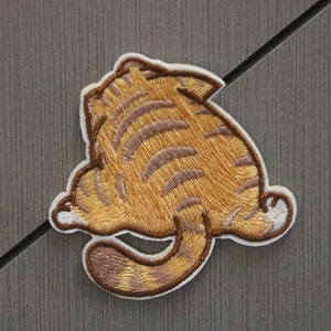 May include: Embroidered patch showing a cartoon cat from behind. The patch has a white border and depicts the cat's back, tail, and hind legs in shades of orange and brown. The patch is designed for application on clothing or accessories.