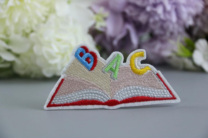 K&ouml;nnte beinhalten: Ein gestickter B&uuml;geleisen-Aufn&auml;her in Form eines offenen Buches mit den Buchstaben "ABC" auf den Seiten. Die Buchstaben sind in verschiedenen Farben: blau, gr&uuml;n und gelb.