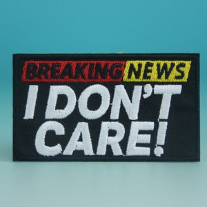 Pode incluir: Etiqueta bordada preta com texto branco que diz "BREAKING NEWS I DON'T CARE!" O texto está em uma fonte em negrito e de bloco. A etiqueta tem uma faixa vermelha e amarela na parte superior.