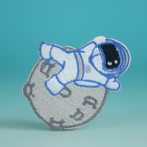 May include: A white and blue embroidered patch of an astronaut in a spacesuit floating on the moon.