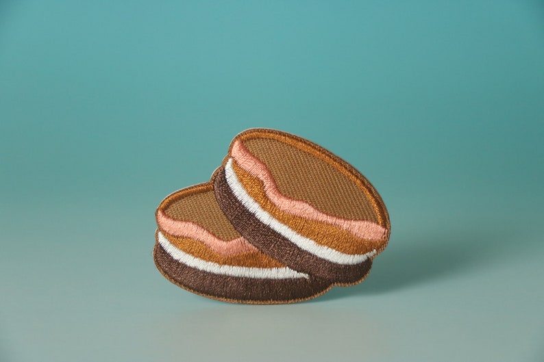 May include: Two embroidered brown and white cookies with pink frosting. The cookies are stacked on top of each other.
