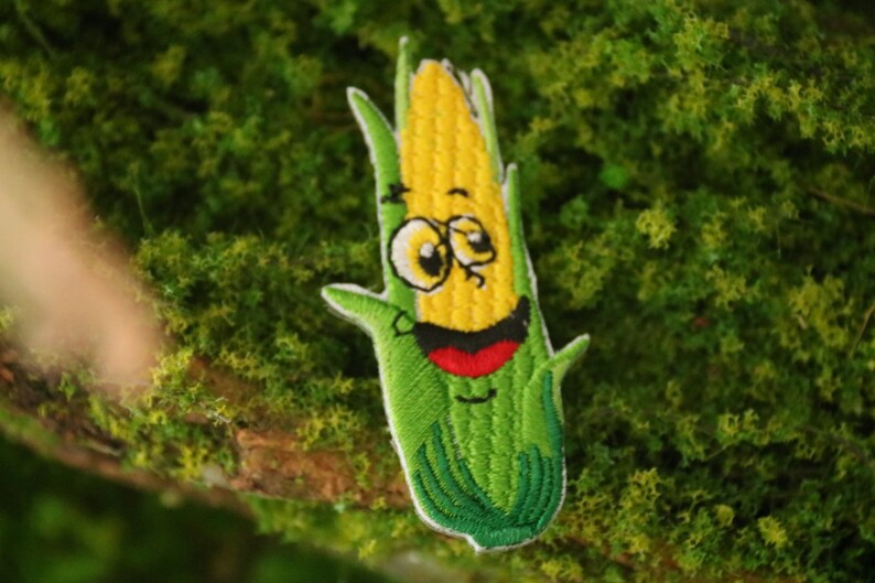 Peut inclure: Un patch brod&eacute; repr&eacute;sentant un &eacute;pi de ma&iuml;s avec un visage souriant. L'&eacute;pi de ma&iuml;s est vert et jaune avec un contour blanc et est entour&eacute; de feuilles vertes.