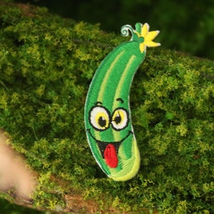 May include: Green embroidered patch of a smiling cucumber with a red tongue and yellow flower on top.