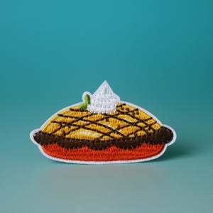 Puede incluir: Un parche bordado que representa una tarta con una cucharada de crema batida en la parte superior. La tarta es amarilla y marrón, y el parche está sobre un fondo blanco.