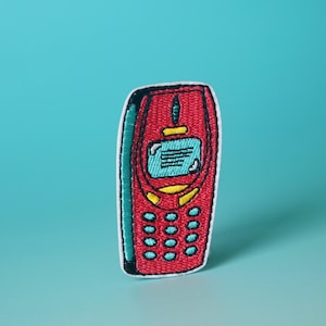 May include: An embroidered patch of a vintage mobile phone, featuring a red body, blue screen and keypad.