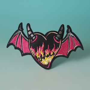 Könnte beinhalten: Schwarzer herzförmiger Patch mit roten und gelben Flammen und schwarzen Teufelshörnern. Das Herz hat rosa Flügel.