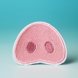 Op de afbeelding: Een roze varkensneus patch met witte stiksels rond de randen. De patch heeft twee roze neusgaten.