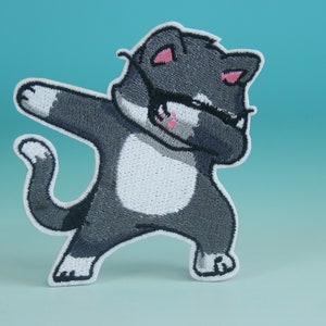 Könnte beinhalten: Gestickter Bügelaufkleber mit einem grauen und weißen Kätzchen, das den Dab macht. Die Katze hat rosa Ohren und eine schwarze Maske.