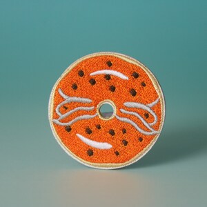 Puede incluir: Parche bordado naranja con forma de donut con detalles blancos y negros.