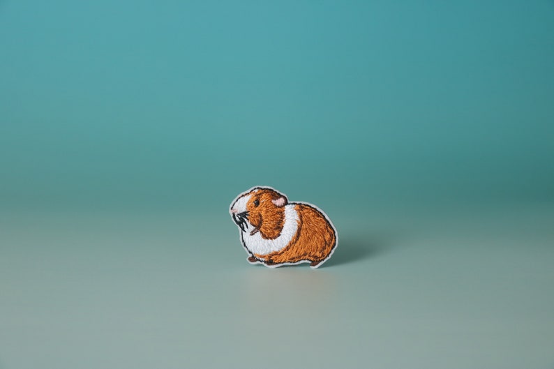 K&ouml;nnte beinhalten: Ein gestickter Aufn&auml;her mit einem braunen und wei&szlig;en Meerschweinchen. Das Meerschweinchen blickt nach links und hat einen wei&szlig;en Fleck um den Hals.