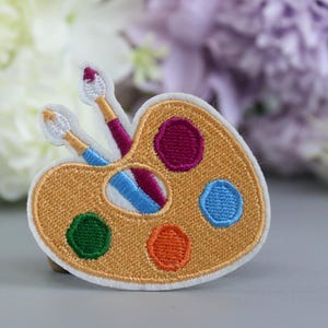 May include: A white embroidered patch featuring a yellow paint palette with two paintbrushes. The palette has five paint wells, filled with red, blue, green, orange, and purple paint.