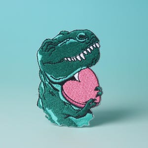 Pode incluir: Dinossauro verde bordado segurando um objeto rosa em forma de cora&ccedil;&atilde;o.