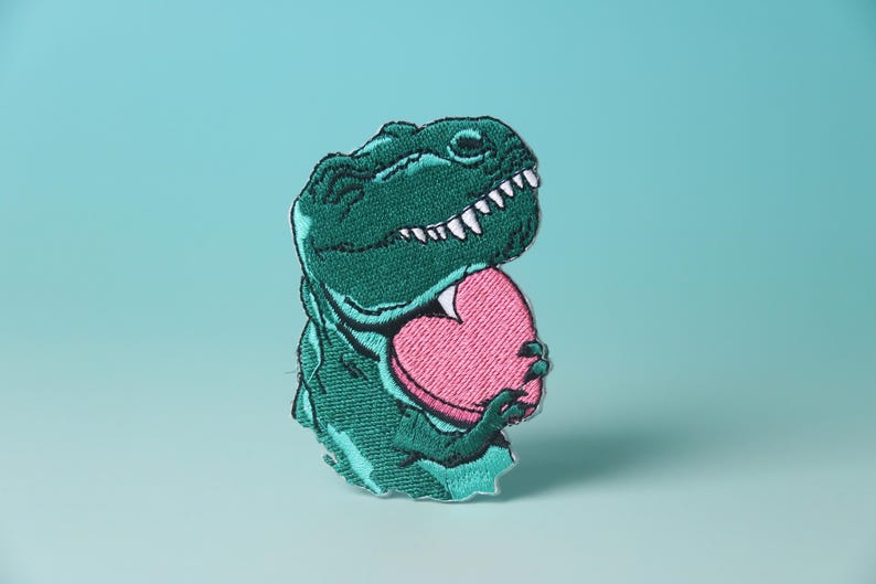 Pode incluir: Dinossauro verde bordado segurando um objeto rosa em forma de cora&ccedil;&atilde;o.
