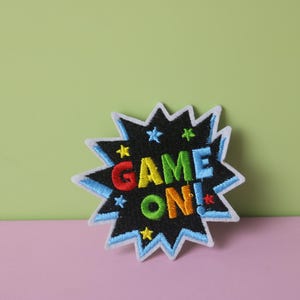 May include: A colourful embroidered patch with a white outline in the shape of a starburst. The patch says "GAME ON!" in rainbow colours.