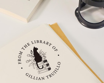 Może przedstawiać: Czarny stempel z tuszem z napisem "FROM THE LIBRARY OF GILLIAN TRUJILLO" otaczającym ilustrację kota na książkach i kwiatach. Stempel znajduje się na białej książce, z drugą książką widoczną w tle.