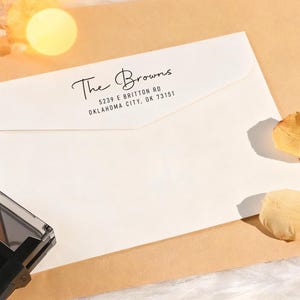 May include: A white envelope with the address "The Browns, 5239 E Britton Rd, Oklahoma City, OK 73151" printed in black script. The envelope is on a tan surface with dried flower petals and a stamp.