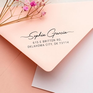 Op de afbeelding: Een lichtroze envelop met de naam "Sophia Garcia" en adresgegevens in het zwart gedrukt. Delicate roze bloemen en stelen sieren de envelop. De envelop is gedeeltelijk open en onthult een witte binnenkant.