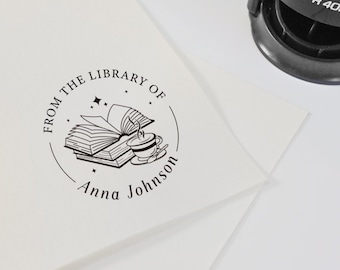 Może przedstawiać: Czarny stempel samotuszujący na białej powierzchni. Stempel ma okrągły wzór z napisem "From the Library of Anna Johnson" otaczającym ilustrację książek i filiżanki do herbaty. Częściowo widoczna jest biała koperta.