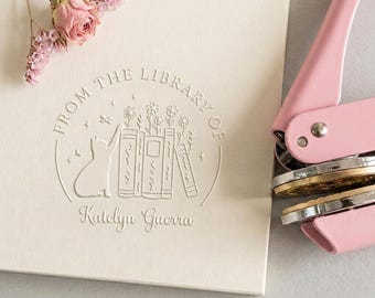 Może przedstawiać: Personalizowana sucha pieczęć z różowym uchwytem i okrągłą metalową matrycą. Wytłoczenie na kremowym papierze przedstawia kota sięgającego do książek z kwiatami, napis "FROM THE LIBRARY OF" oraz imię "Katelyn Guerra" obok suszonej róży.
