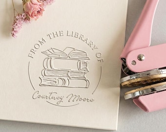 Może przedstawiać: Jasnoróżowe metalowe narzędzie do tłoczenia obok kremowego papieru z okrągłym tłoczonym wzorem stosu książek z napisem FROM THE LIBRARY OF Courtney Moore oraz małą gałązką suszonych różowych kwiatów umieszczoną w lewym górnym rogu.