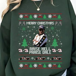 May include: Dark green sweatshirt with a festive Christmas design. The design includes the text "MERRY CHRISTMAS", snowflakes, race cars, and the phrase "RAISE HELL PRAISE DALE". A portrait of a man wearing sunglasses is also featured.