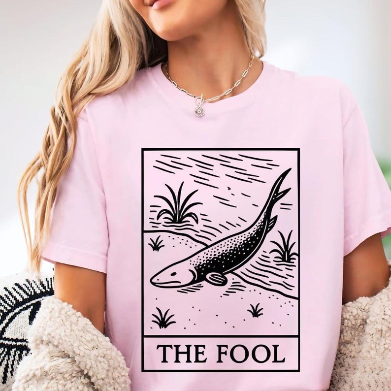 A woman wearing a pink t-shirt with a graphic design of a fish and the text "THE FOOL" printed on it.