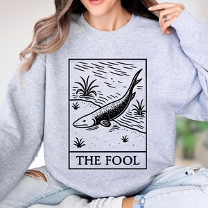 A woman wearing a grey sweatshirt with a graphic design depicting a fish and the text "THE FOL" printed on it.